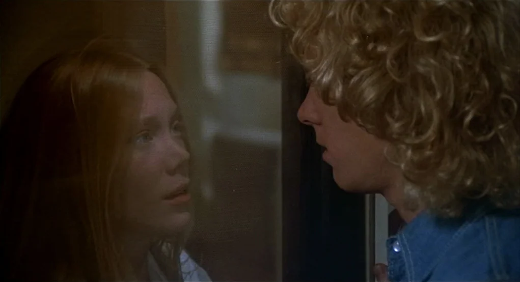 Carrie (1976) movie still: medium — A dimly lit indoor space with a dark frame separating the two subjects. A young man with blonde curls stands…