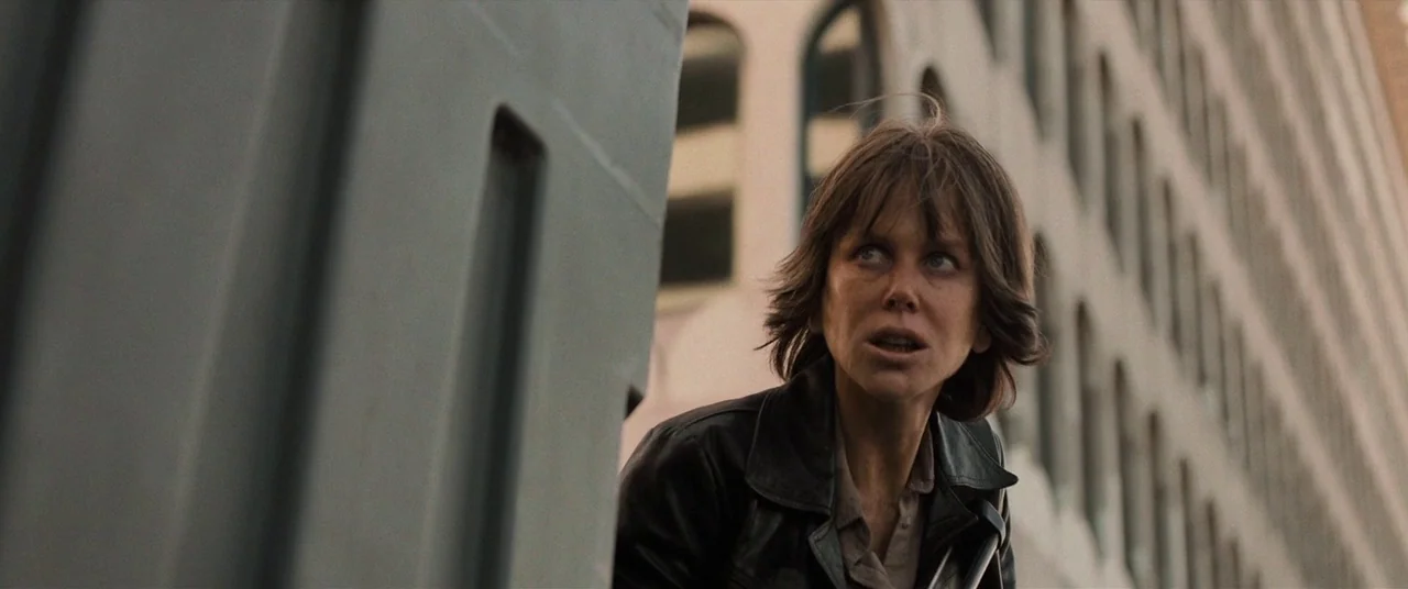 Destroyer (2018) movie still: medium — A woman peeks around the edge of a grey pillar on a metropolitan street. Tall buildings with arched windows…