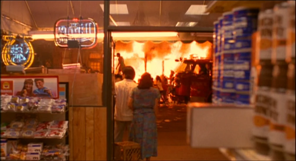 Do The Right Thing (1989) movie still: wide — A convenience store interior looking out at a building engulfed in orange flames. A man and woman…