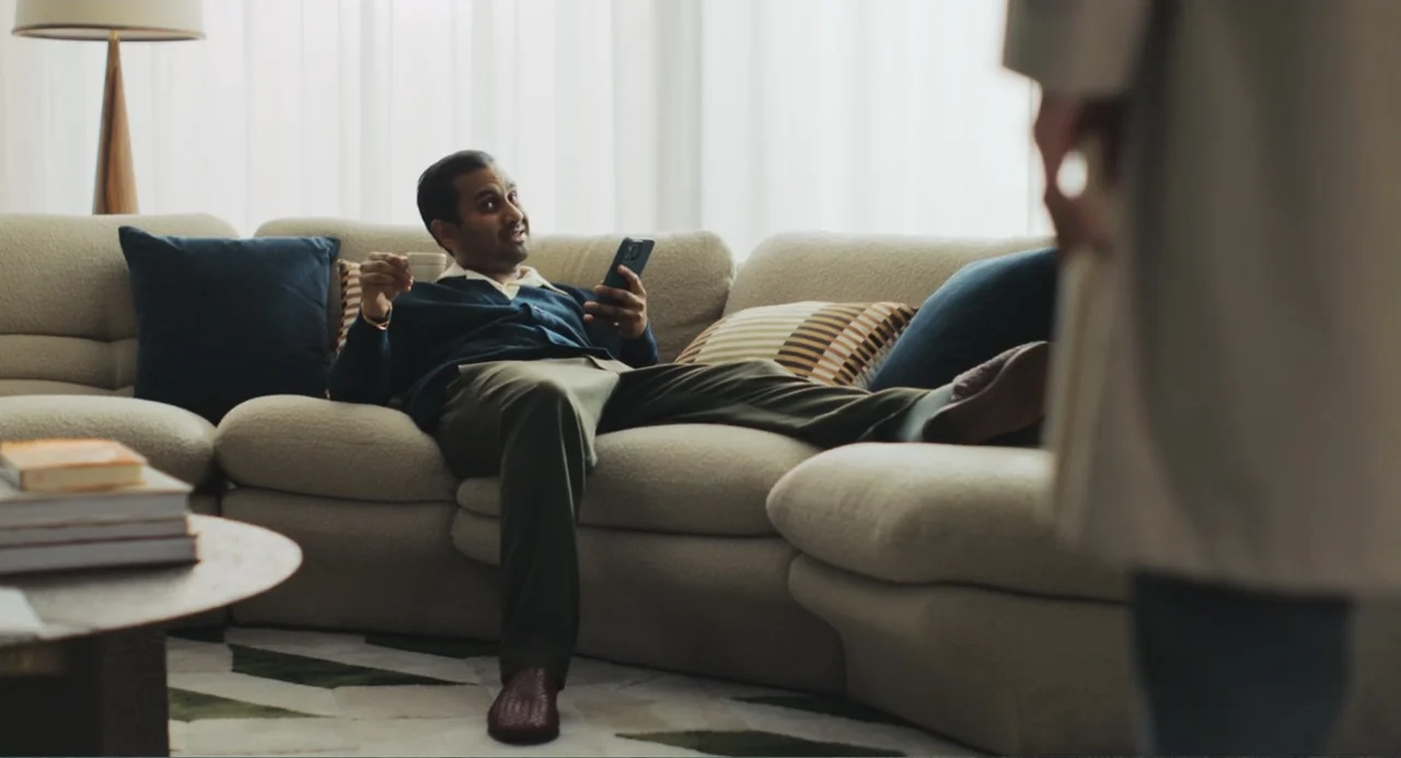 Good Fortune (2025) movie still: medium — A living room with a large light-colored sectional sofa and sheer window curtains. A man sits on the sofa…