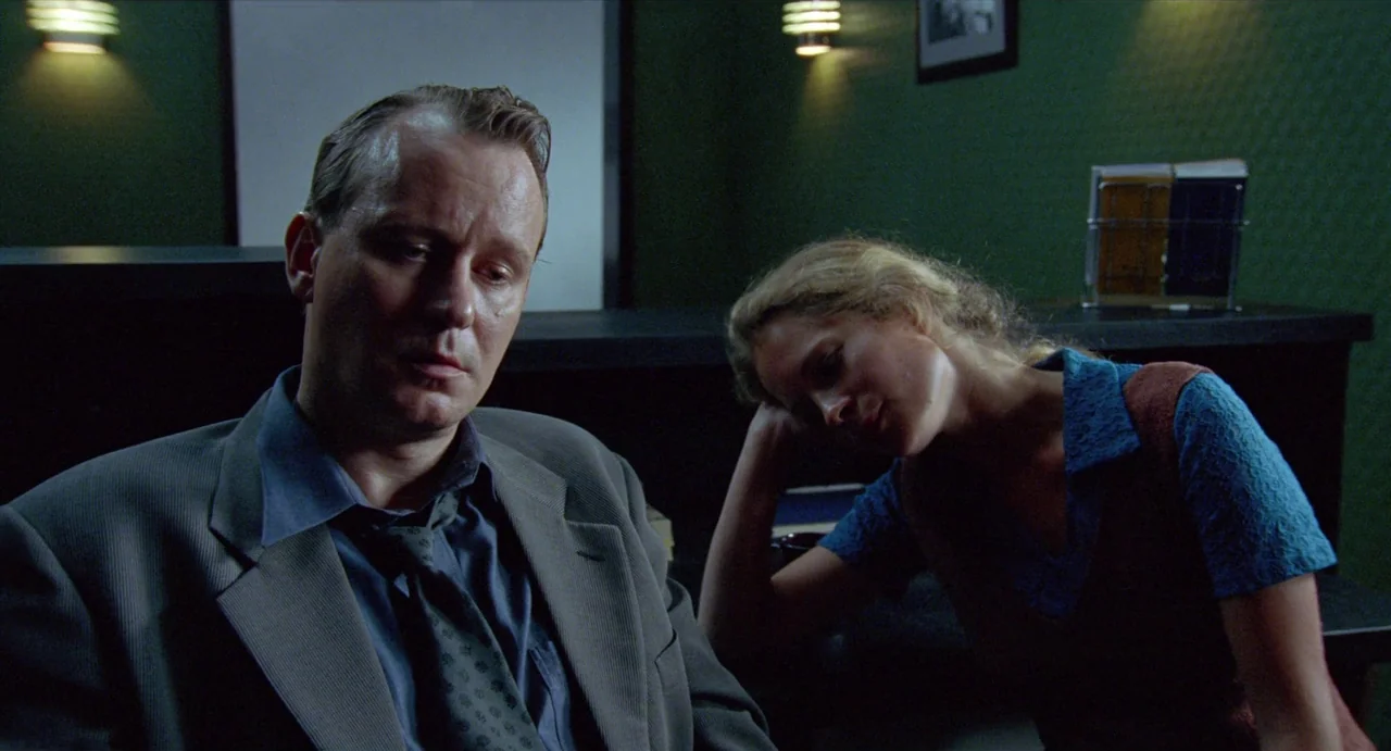 Insomnia (1997) movie still: medium — A dimly lit room features walls painted dark green with wall-mounted lamps. A man in a suit and a woman in a…