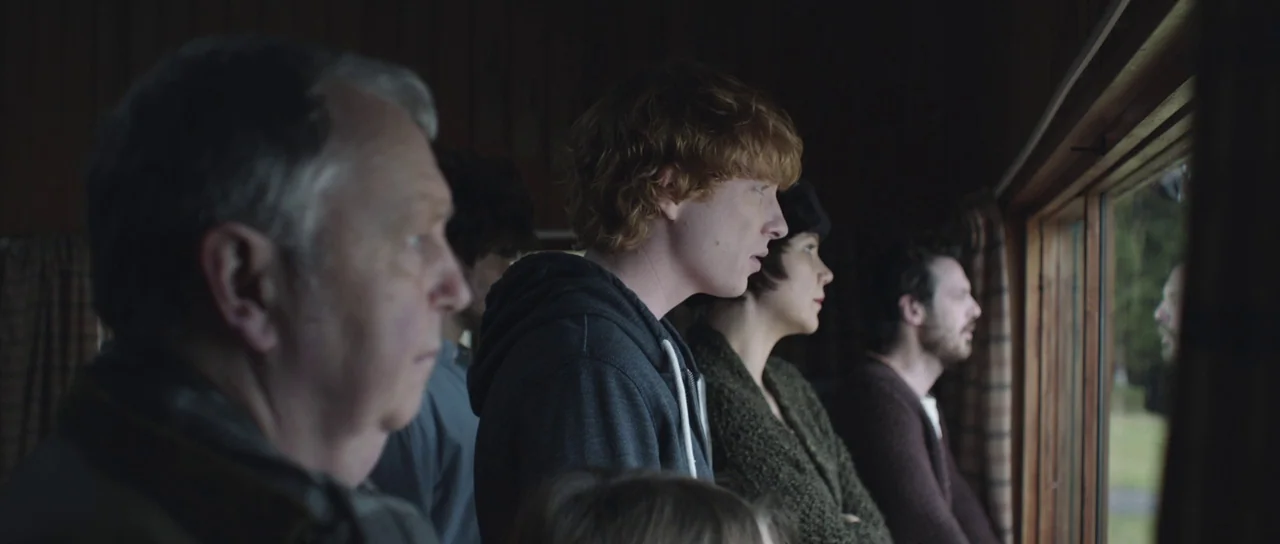 Frank (2014) movie still: medium — A dimly lit wooden interior with a long rectangular window overlooking an outdoor area. Several people stand in a…