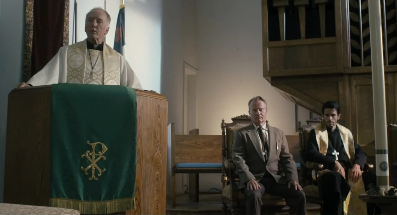 Gates of Darkness (2019) movie still: medium — A church altar features a wooden pulpit draped in a green cloth with gold lettering. Three men sit or…