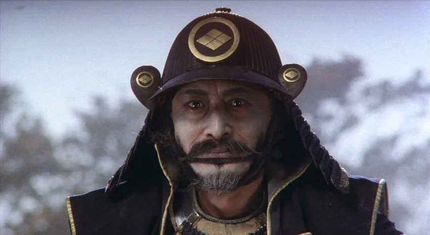Kagemusha (1980) movie still: closeup — An outdoor location with a blurred treeline visible in the bright background. A man wearing a dark samurai…
