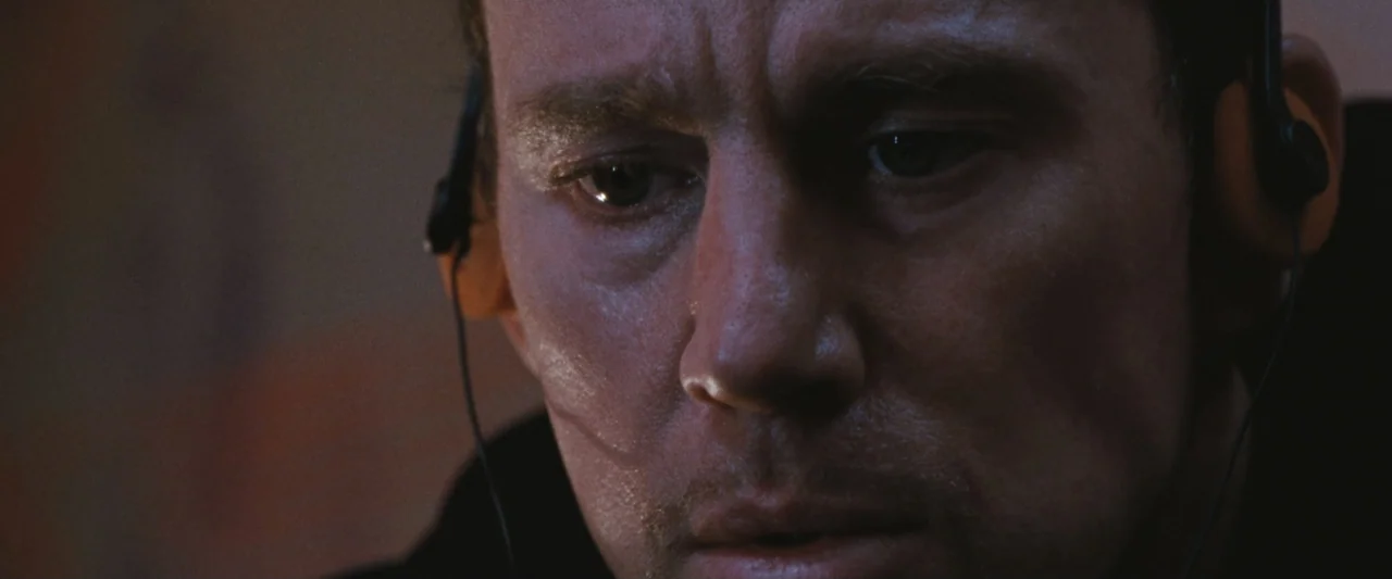 Roofman (2025) movie still: closeup — A dark indoor setting with minimal detail visible in the background. A man wearing a headset looks down toward…