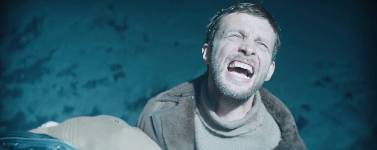 Petrov’s Flu (2021) movie still: closeup — A dark cave wall with rough textures serves as the backdrop. A man in a high-neck sweater is screaming…