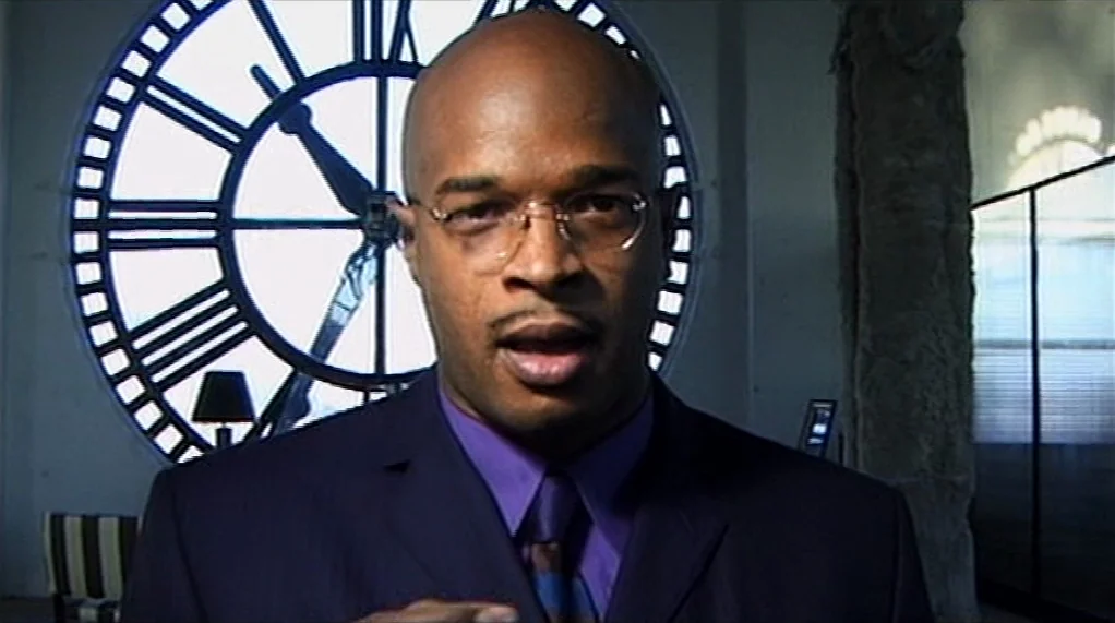 Bamboozled (2000) movie still: closeup — An interior space features a massive structural clock face set into a large window. A man in a suit stands…