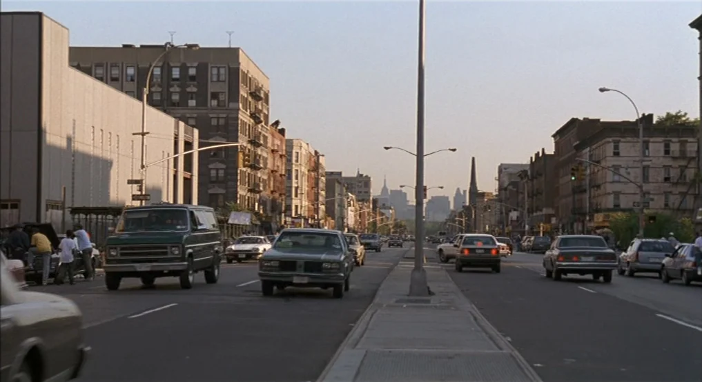 Light Sleeper (1992) movie still: establishing — A wide city street is lined with multi-story apartment buildings and parked cars. Traffic flows…