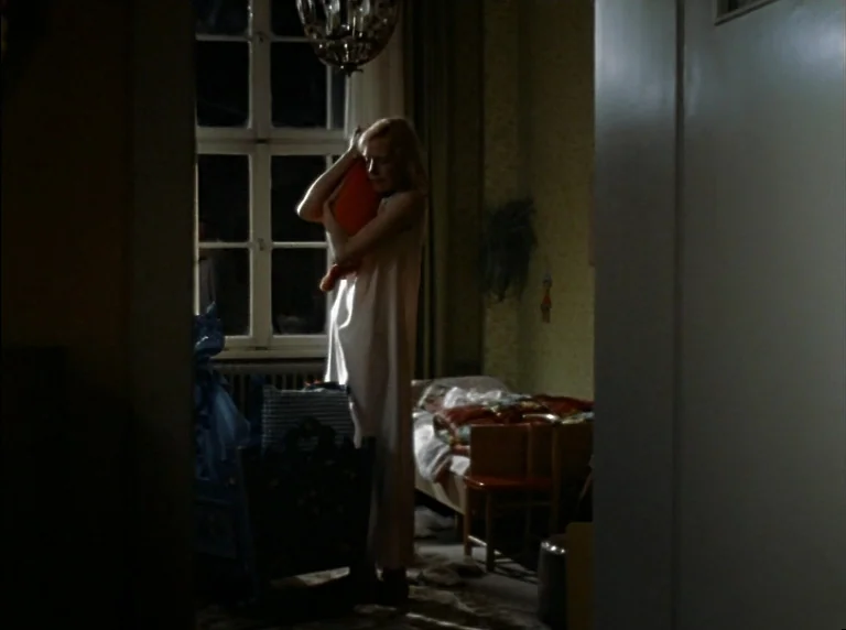 Fear Of Fear (1975) movie still: fullbody — A dark nursery room features a wooden crib and an unmade bed in the corner. A woman stands by a window…
