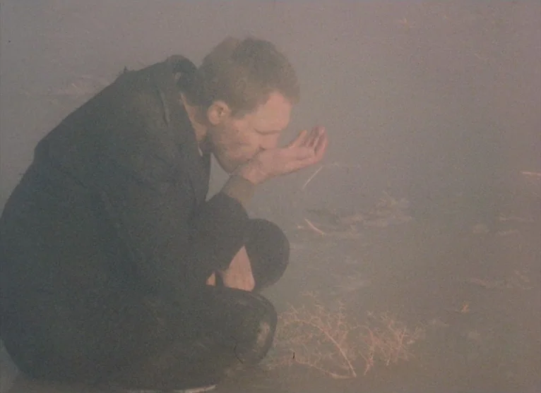 A Visitor to a Museum (1989) movie still: medium — An open, hazy area shrouded in thick, gray fog at ground level. A lone man in a dark jacket…
