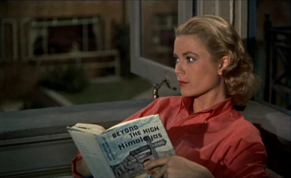 Rear Window (1954) movie still: medium — A woman sits on a balcony at night holding a book titled Beyond the High Himalayas. The blurred background…