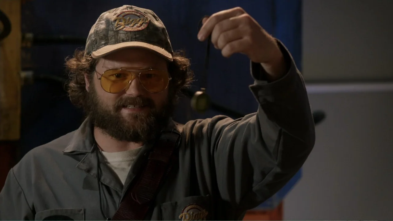 Rufus 2 (2017) movie still: medium — A man stands in an industrial workspace with grey walls and muted equipment. He lifts a small round object on a…