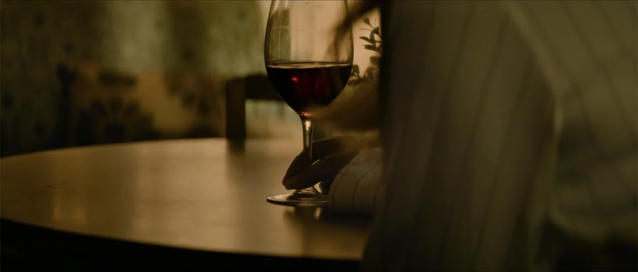 Crescent City (2024) movie still: medium — A dimly lit dining room features a wooden table and a red wine glass. A person seen from the back rests…