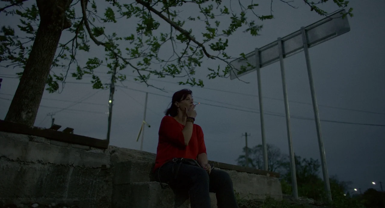 Blackbird Blackbird Blackberry (2023) movie still: medium — A woman sits on a concrete structure under a dark, overcast evening sky. She holds a lit…