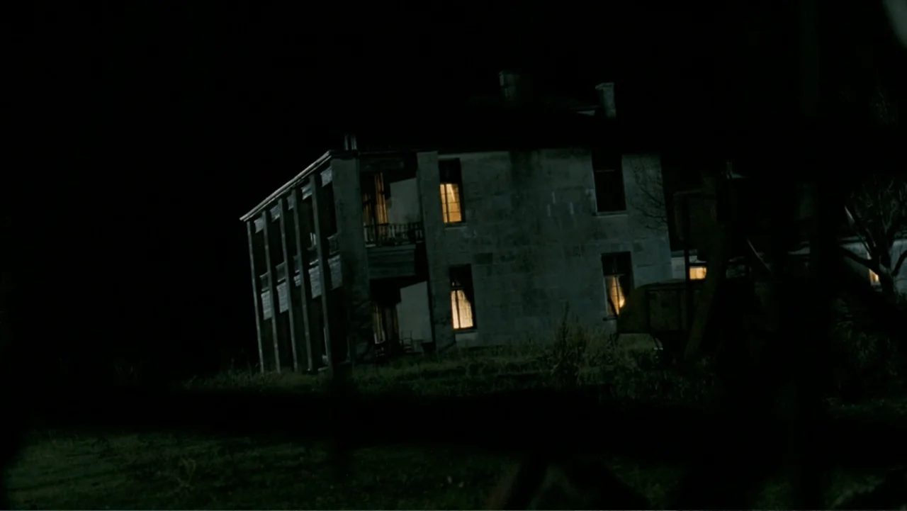 The Texas Chainsaw Massacre: The Beginning (2006) movie still: wide — A two-story stone house sits on a dark grassy hill at night. Warm light spills…