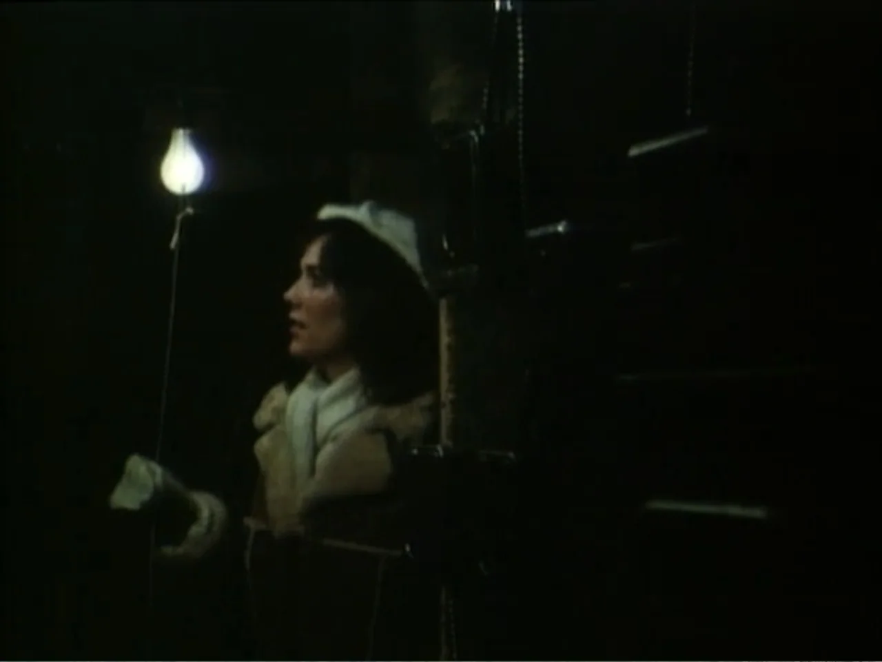 Curtains (1995) movie still: medium — A dark, indistinct interior is illuminated by a single low-hanging light bulb. A young woman in a beret and…
