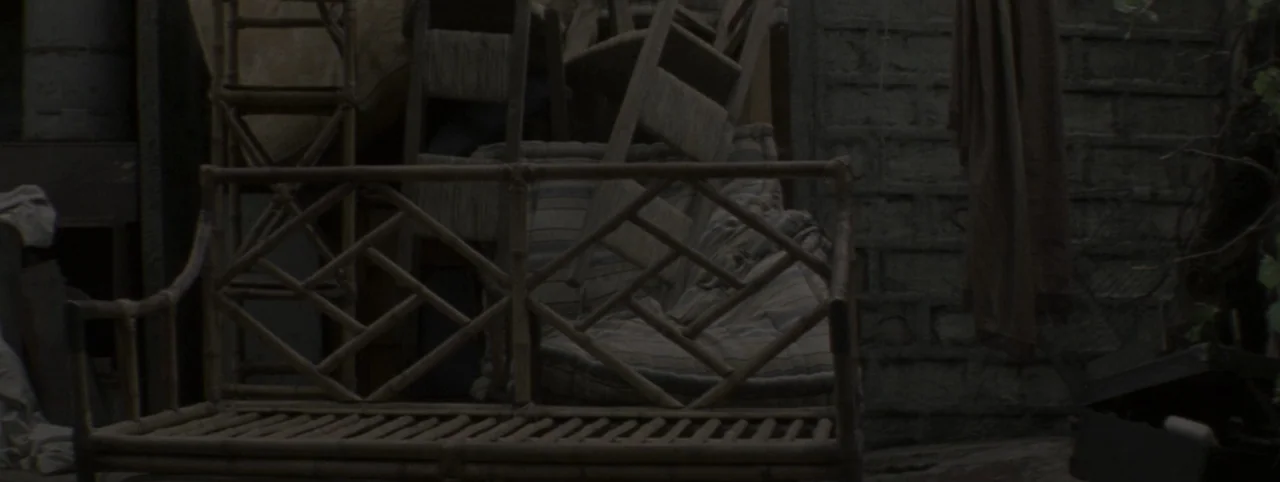 Post Mortem (2010) movie still: medium — An interior space contains a cluster of aged, discarded wooden and bamboo furniture items. The dark area is…