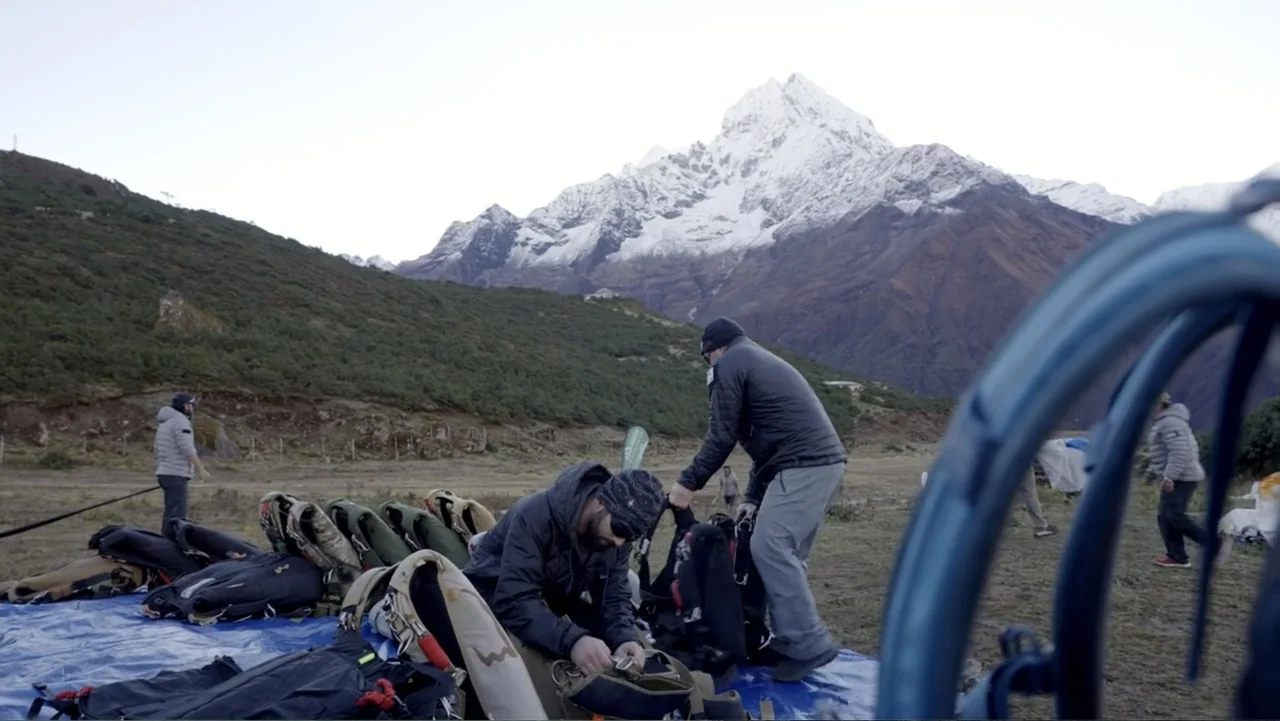 Drop Zone Everest (2023) movie still: wide — A high-altitude base camp sits in a valley beneath a snow-capped mountain peak. Several men in outdoor…