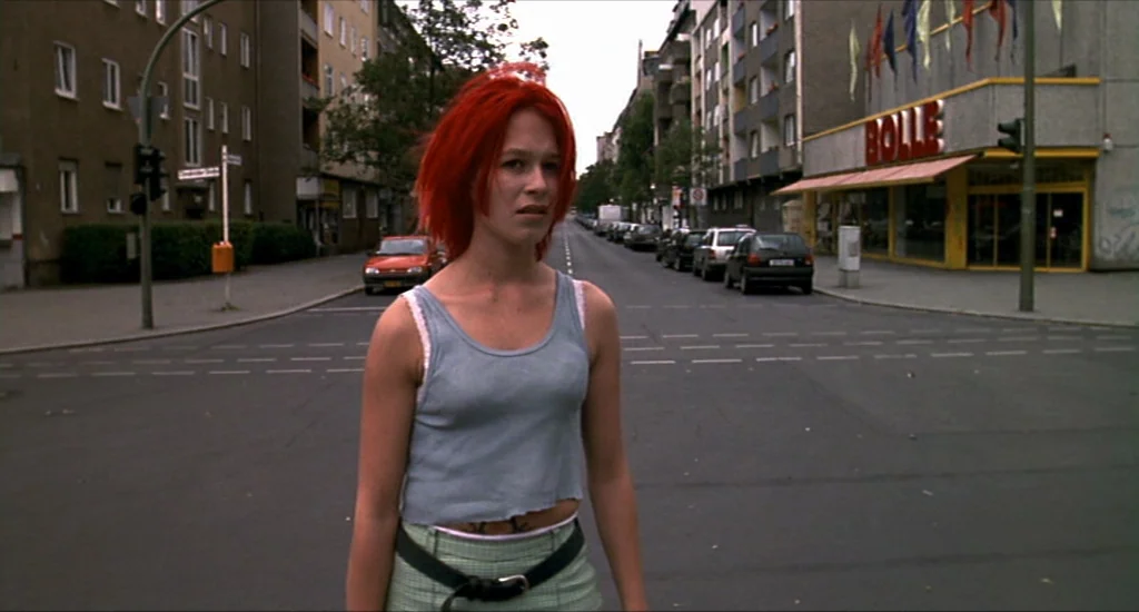 Run Lola Run (1998) movie still: medium — A woman stands in the center of an empty intersection in a city. Buildings line the street with a grocery…