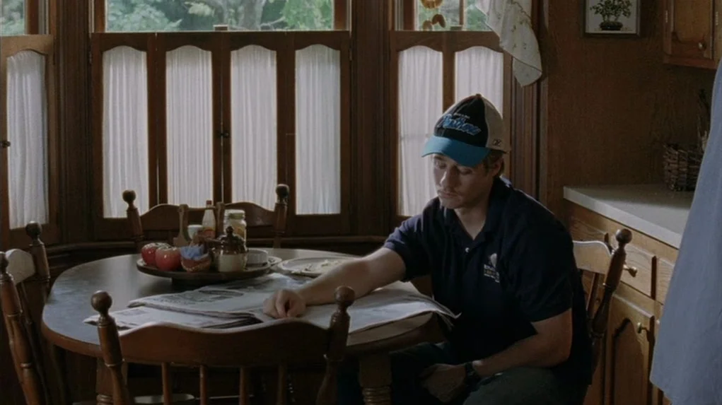 Junebug (2005) movie still: medium — A wooden dining nook features a circular table with a newspaper and apples. A young man sits in a chair reading…