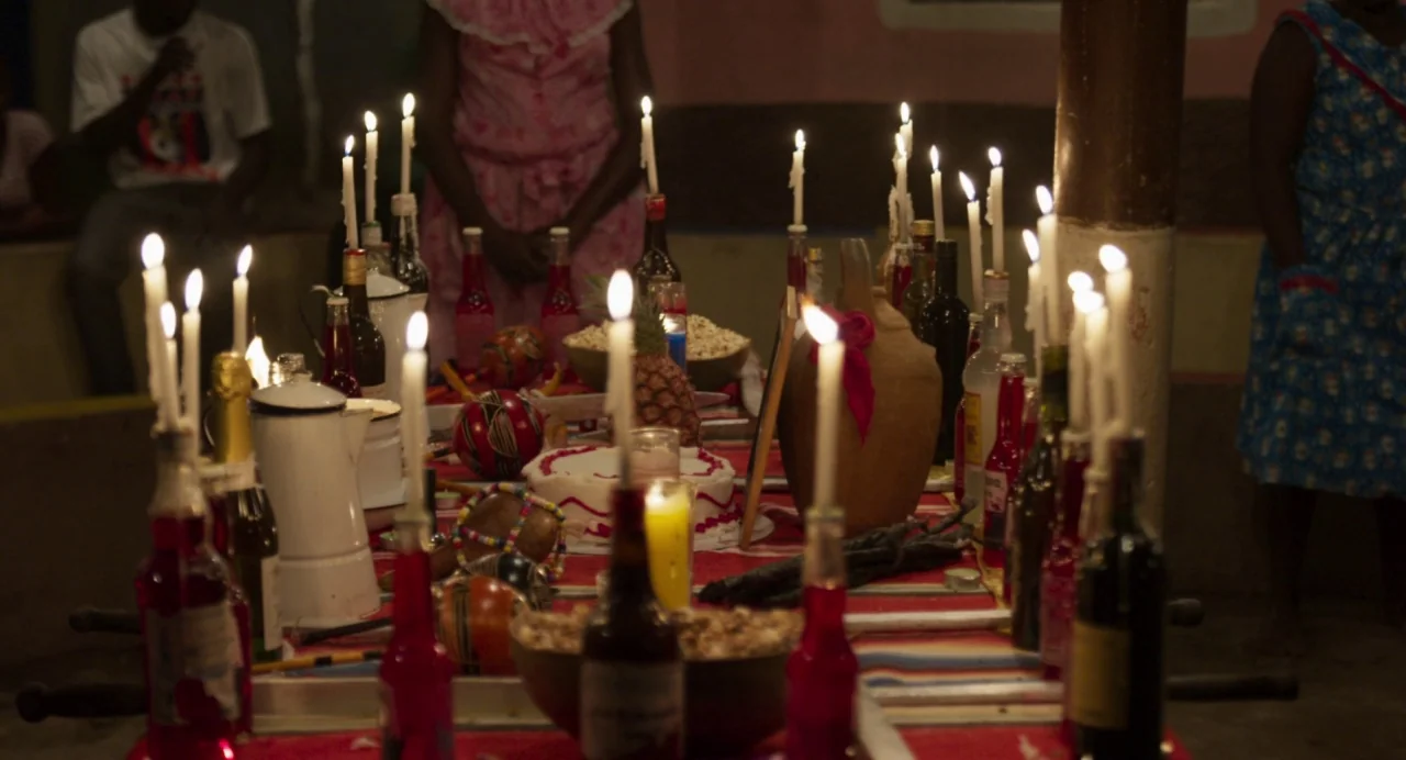 Zombi Child (2019) movie still: medium — A table is covered with a patterned red cloth and adorned with many candles. Numerous bottles, a cake, and…
