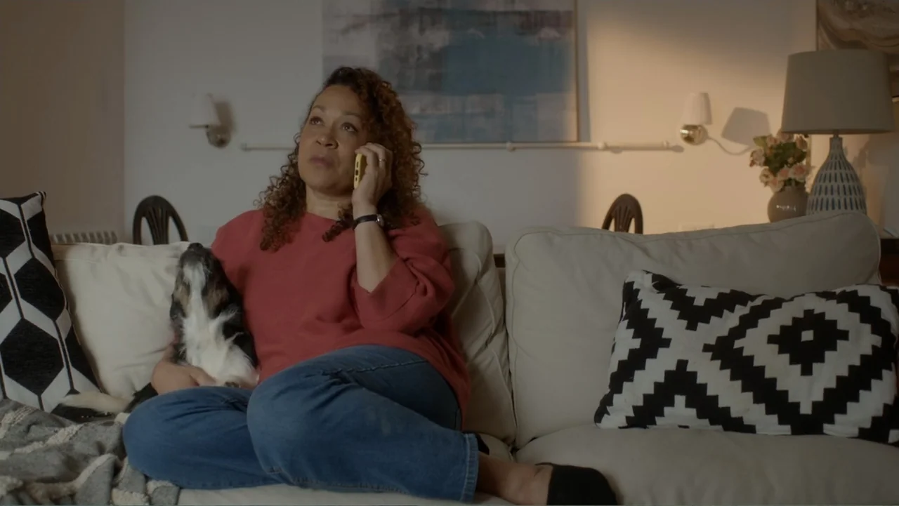 A Pinch of Portugal (2023) movie still: medium — A living room interior features a white sofa, a table lamp, and framed art. A woman sits on the…