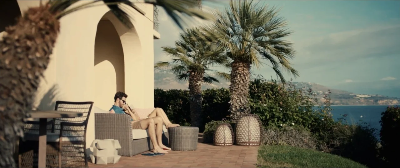 Plus One (2019) movie still: wide — A sunny outdoor patio overlooks a coastline with palm trees and calm blue water. Two people sit side by side on…