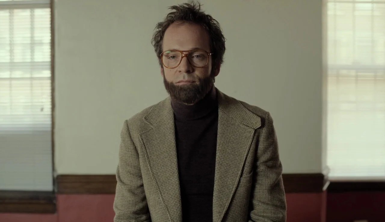 Experimenter (2015) movie still: medium — A man stands centered in a sparsely furnished room with light-colored walls. He looks directly at the…