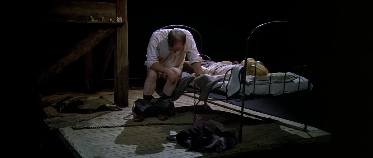 Dogville (2003) movie still: wide — A minimalist set featuring a metal bed frame on a raised wooden platform. A man in a white shirt sits on the…
