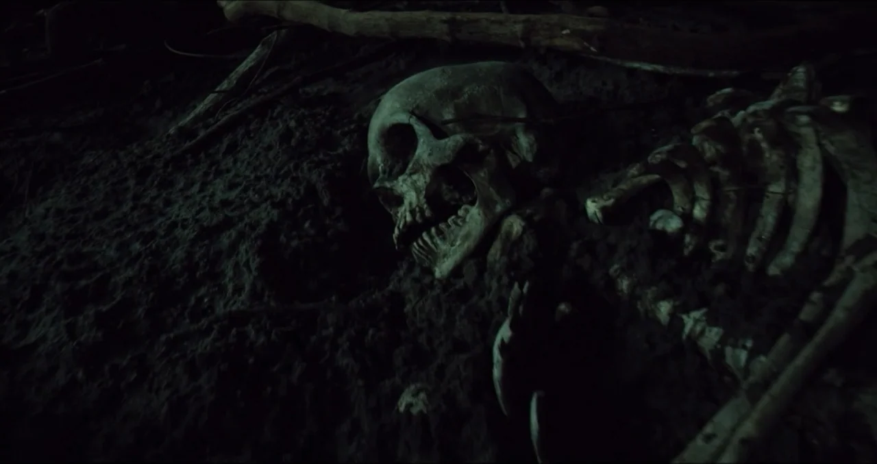 Eye for an Eye (2025) movie still: closeup — A partially unearthed human skeleton rests in a shallow depression in dark soil. Twigs and loose…