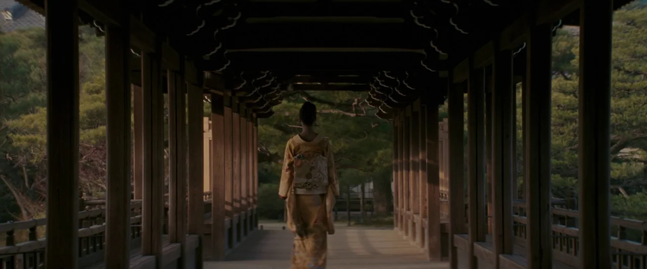Memoirs of a Geisha (2005) movie still: fullbody — A wooden covered walkway leads through a Japanese-style garden with pine trees visible outside. A…