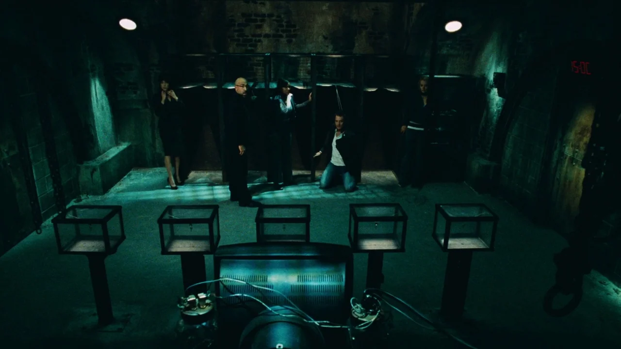 Saw V (2008) movie still: wide — A dark subterranean chamber features exposed brick walls and metal cage-like partitions. Five people stand or kneel…
