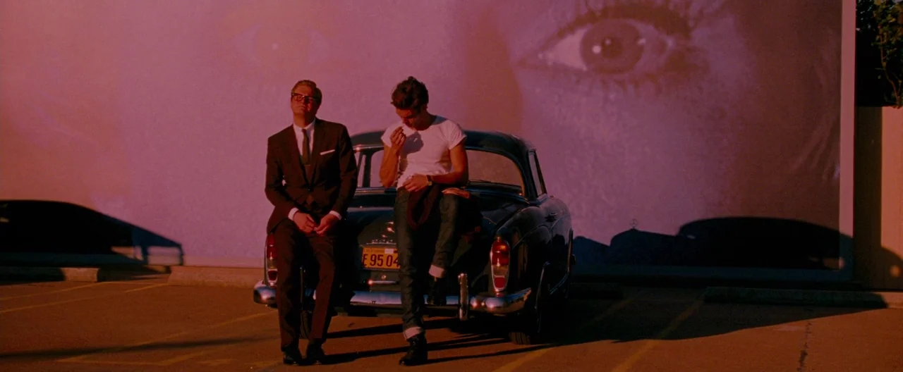 A Single Man (2009) movie still: wide — A dark parking lot at night sits in front of a giant cinema projection screen. Two men sit on the back of a…