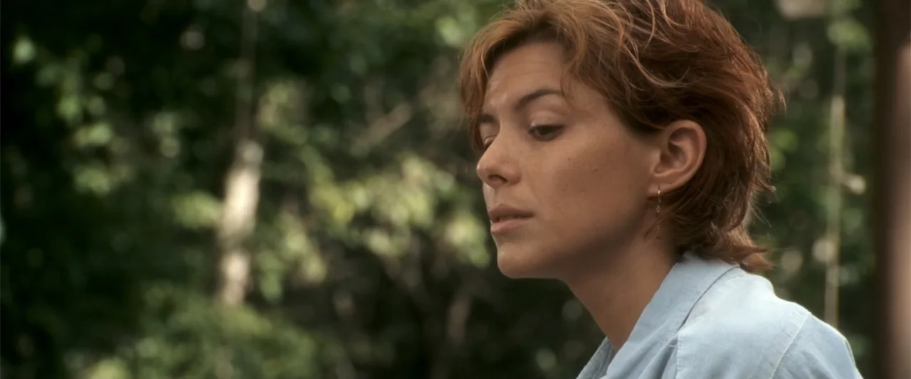 Anaconda (2025) movie still: closeup — A woman stands outdoors in front of a blurry background of green trees. She looks off-camera to the left with…