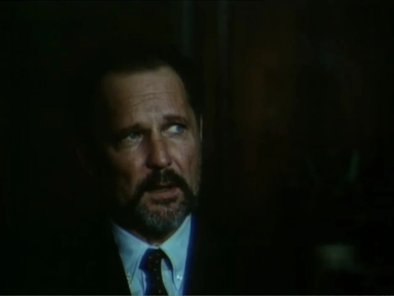 Curtains (1995) movie still: closeup — A man sits in a dimly lit, dark office environment with obscure background details. The subject is angled…