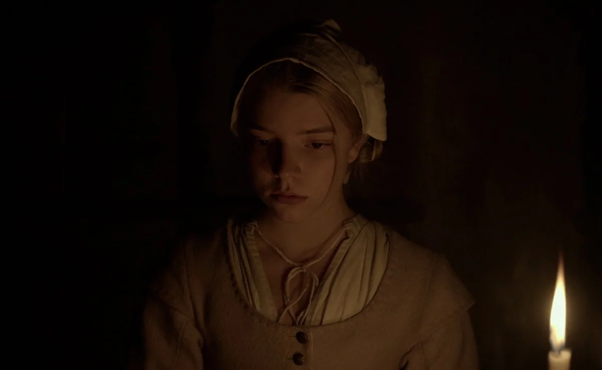 The Witch (2015) movie still: medium — A dimly lit room with stone walls barely visible in the dark background. A young woman with a white cap…