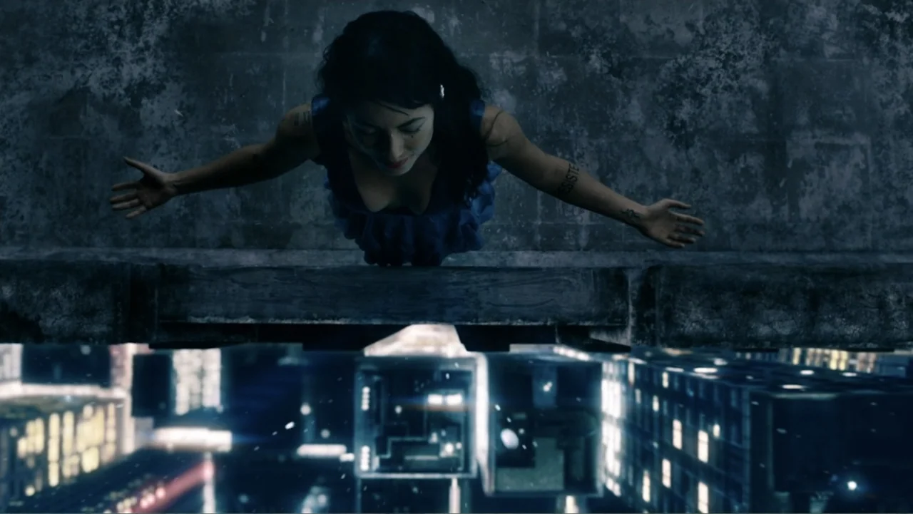 Alice in Marialand (2014) movie still: medium — A woman stands on the edge of a concrete precipice overlooking a city at night. She leans forward…