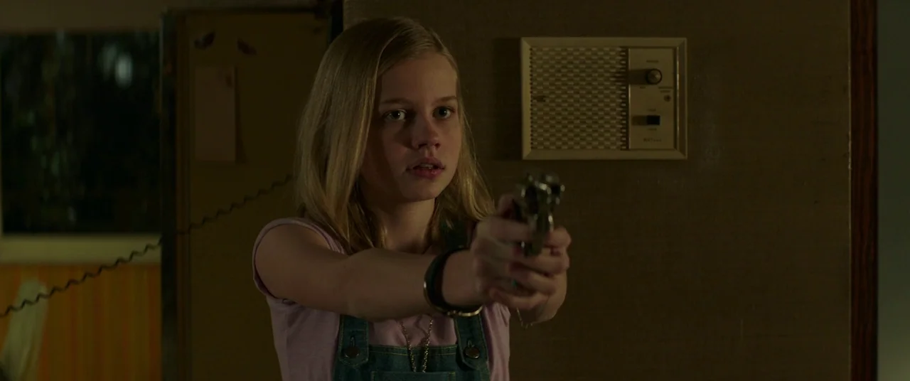 The Nice Guys (2016) movie still: medium — A dark indoor space features a wall-mounted intercom device behind a young girl. The teenage girl stands…