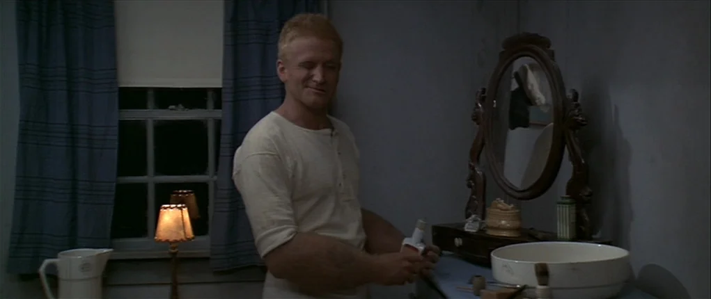Popeye (1980) movie still: medium — A dimly lit room features a wooden mirror and a white porcelain washbasin. A man wearing a light-colored shirt…