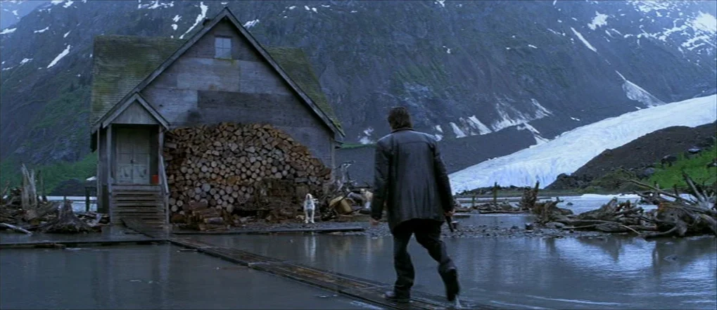 Insomnia (2002) movie still: wide — A rustic wooden cabin sits at the edge of a flooded, glacial mountain valley. A man in a dark coat walks across…
