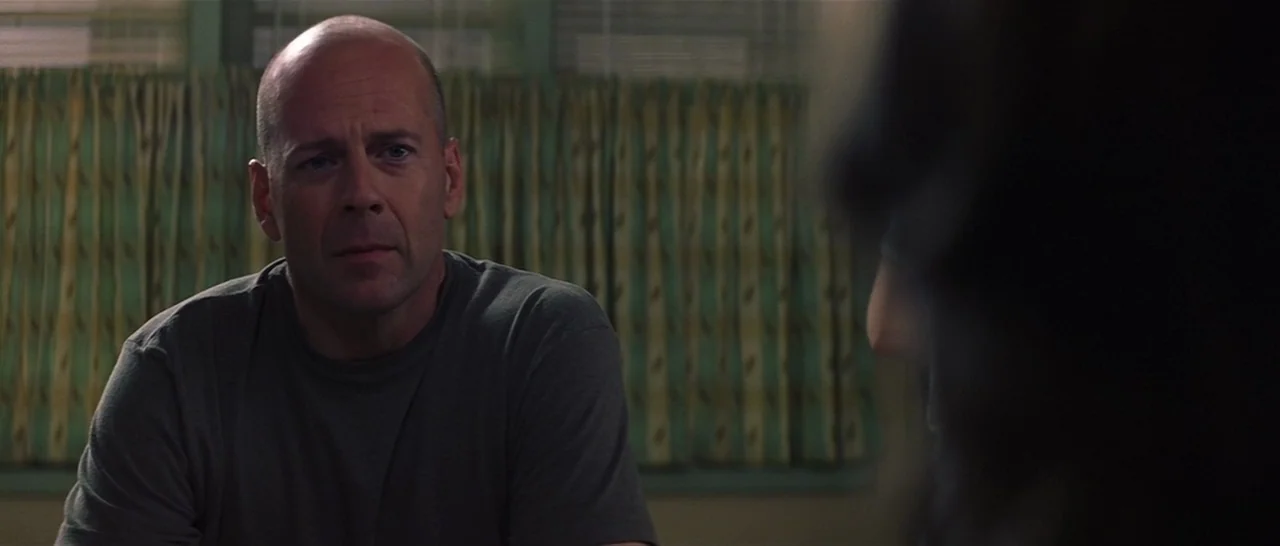 Unbreakable (2000) movie still: medium — A man sits in a dimly lit space with patterned window blinds behind him. The man is looking directly at…
