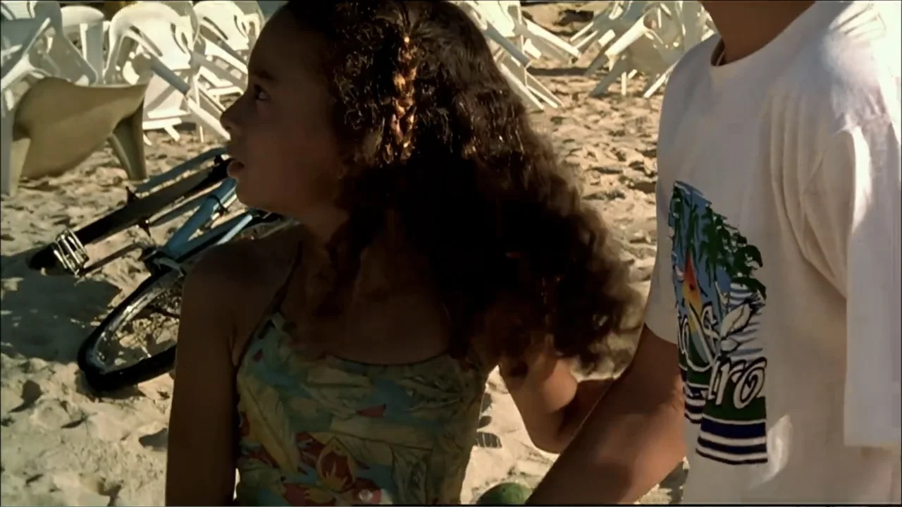 In the Middle of the World (1990) movie still: medium — A sunny sandy beach features scattered plastic chairs and a bicycle on the ground. A young…