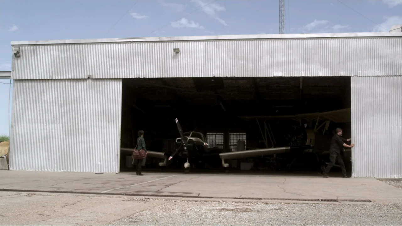 The Messenger (2015) movie still: wide — A metallic corrugated hangar building features a wide opening revealing a light aircraft inside. A man…