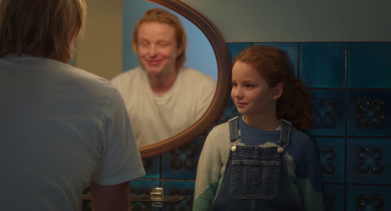 Petite Maman (2021) movie still: medium — A bathroom wall is covered with deep blue patterned square tiles. A young girl in denim overalls stands…