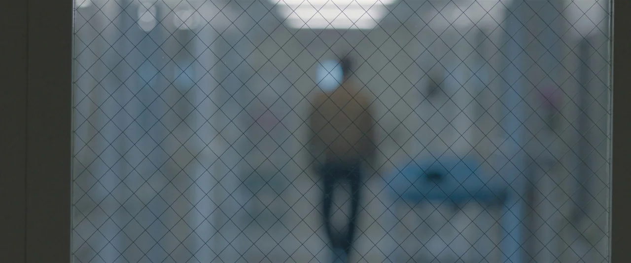 The Gift (2015) movie still: wide — A sterile medical hallway is viewed through a glass panel with a wire grid. A man walks away from the camera…