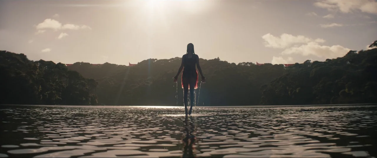 Evil Dead Rise (2023) movie still: wide — A calm lake is framed by densely forested hills under a bright sun. A woman stands in the middle of the…