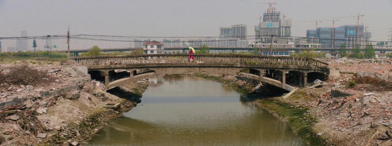 Dead Pigs (2018) movie still: wide — A weathered concrete bridge spans a narrow, murky canal in an area of urban demolition. A lone person with a…