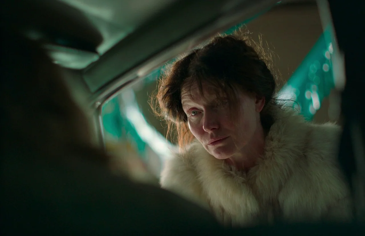 Nitram (2021) movie still: closeup — A woman leans into a car window at night, illuminated by dim ambient light. The interior of the car is dark,…
