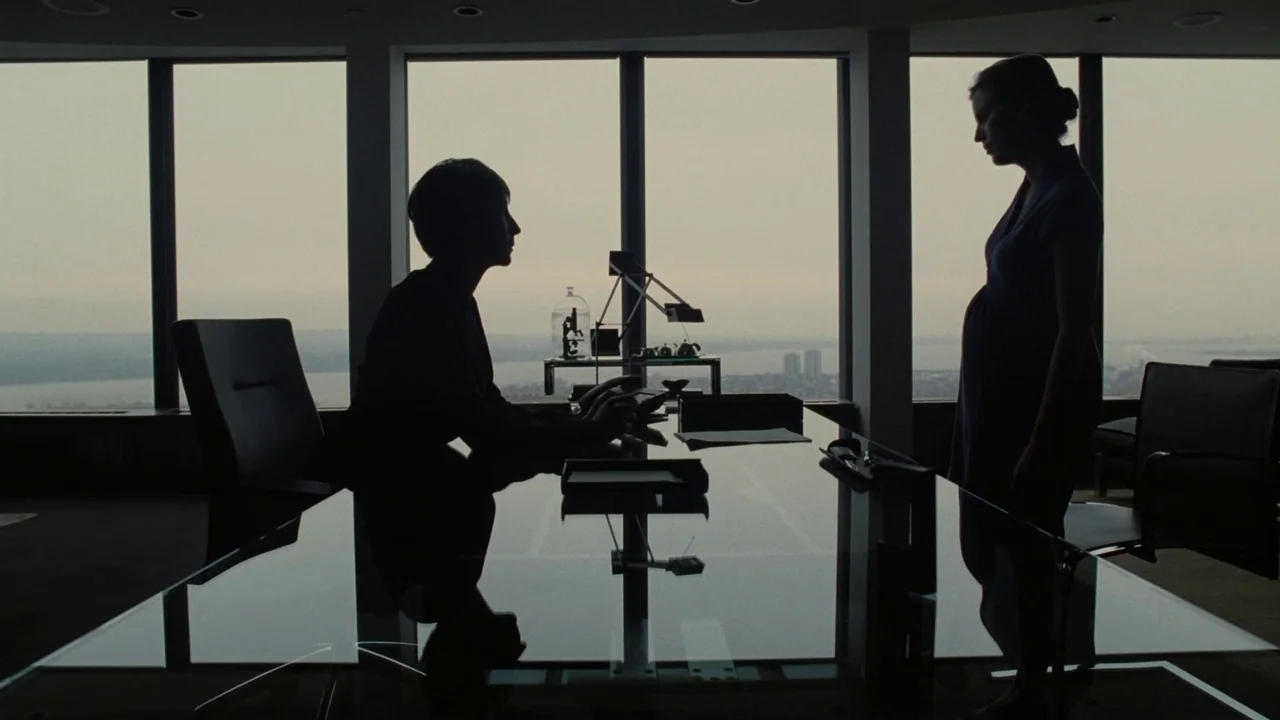 Splice (2009) movie still: wide — A high-rise office with floor-to-ceiling windows looking out over a wide body of water. A seated woman faces a…