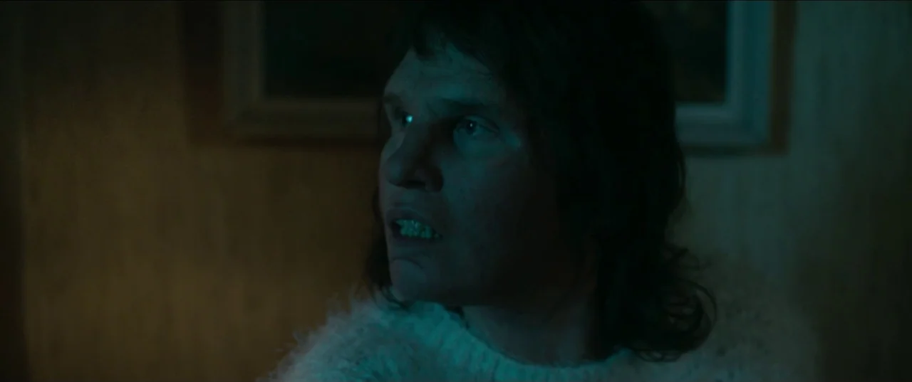 Border movie still: closeup — A dimly lit domestic interior features textured wallpaper and a framed picture on the wall. A man in a white sweater…